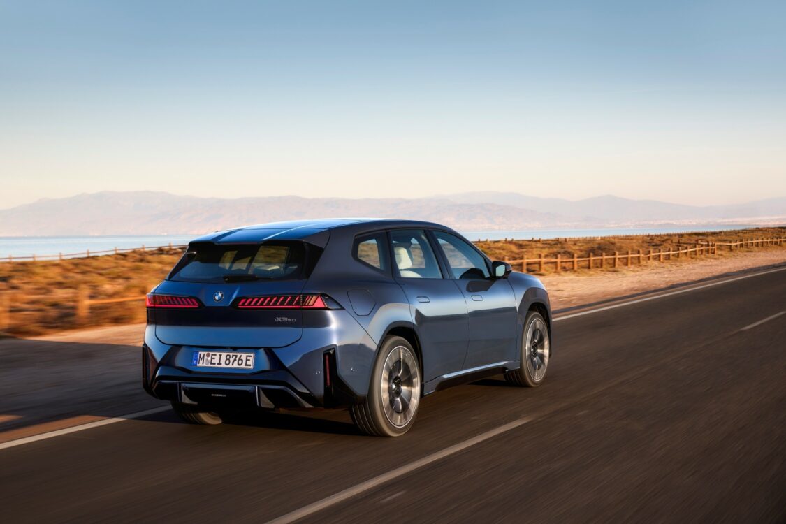 BMW iX3 – design arrière et dynamisme sur route Vue arrière de la BMW iX3 roulant sur une route côtière, avec les feux arrière allumés et un paysage montagneux en arrière-plan.