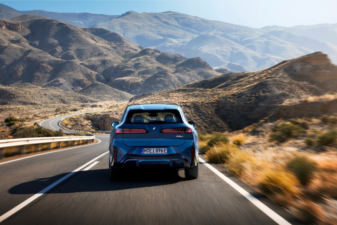 BMW iX3 – équilibre et assurance sur route de montagne Vue arrière de la BMW iX3 roulant sur une route de montagne sinueuse, intégrée dans un paysage minéral et vallonné.