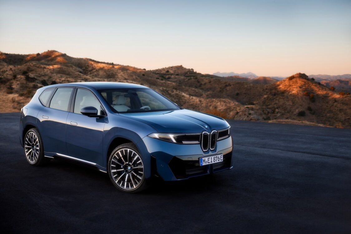 BMW iX3 – présence affirmée et élégance statique BMW iX3 bleue en vue avant trois quarts, stationnée sur une esplanade avec en arrière-plan un paysage montagneux au coucher du soleil.