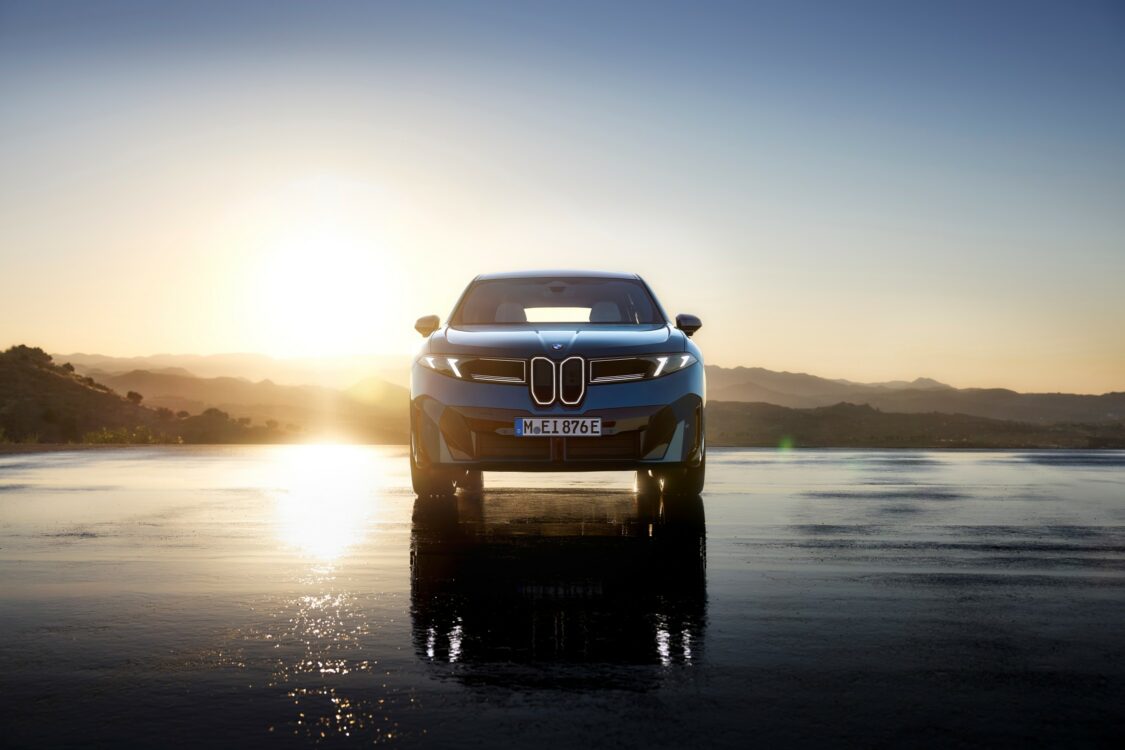 BMW iX3 – silhouette avant au soleil couchant BMW iX3 vue de face, stationnée sur une surface réfléchissante au coucher du soleil, avec les montagnes en arrière-plan.