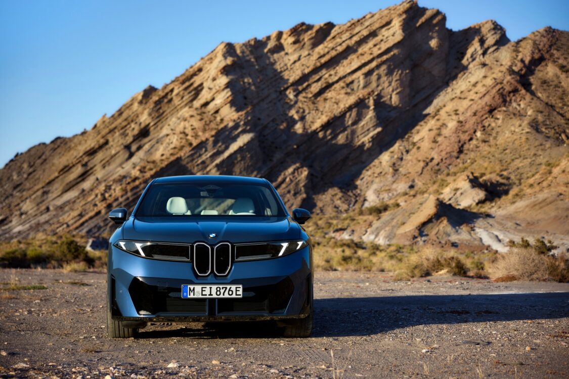 BMW iX3 – puissance face au relief BMW iX3 vue de face, carrosserie bleue, stationnée devant une formation rocheuse inclinée dans un paysage désertique.