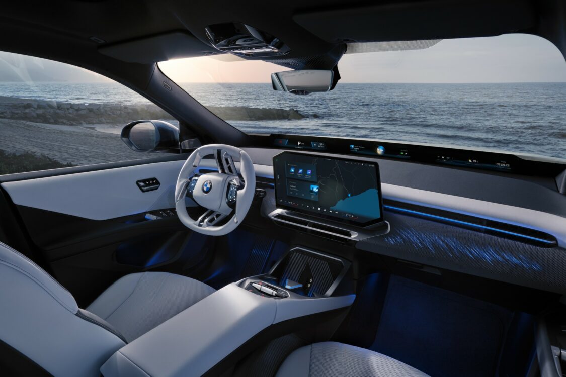 Intérieur BMW iX3 – cockpit avec vue sur la mer Vue intérieure de la BMW iX3 côté conducteur, avec volant blanc, écran central affichant la navigation et décor LED bleuté face à un paysage marin
