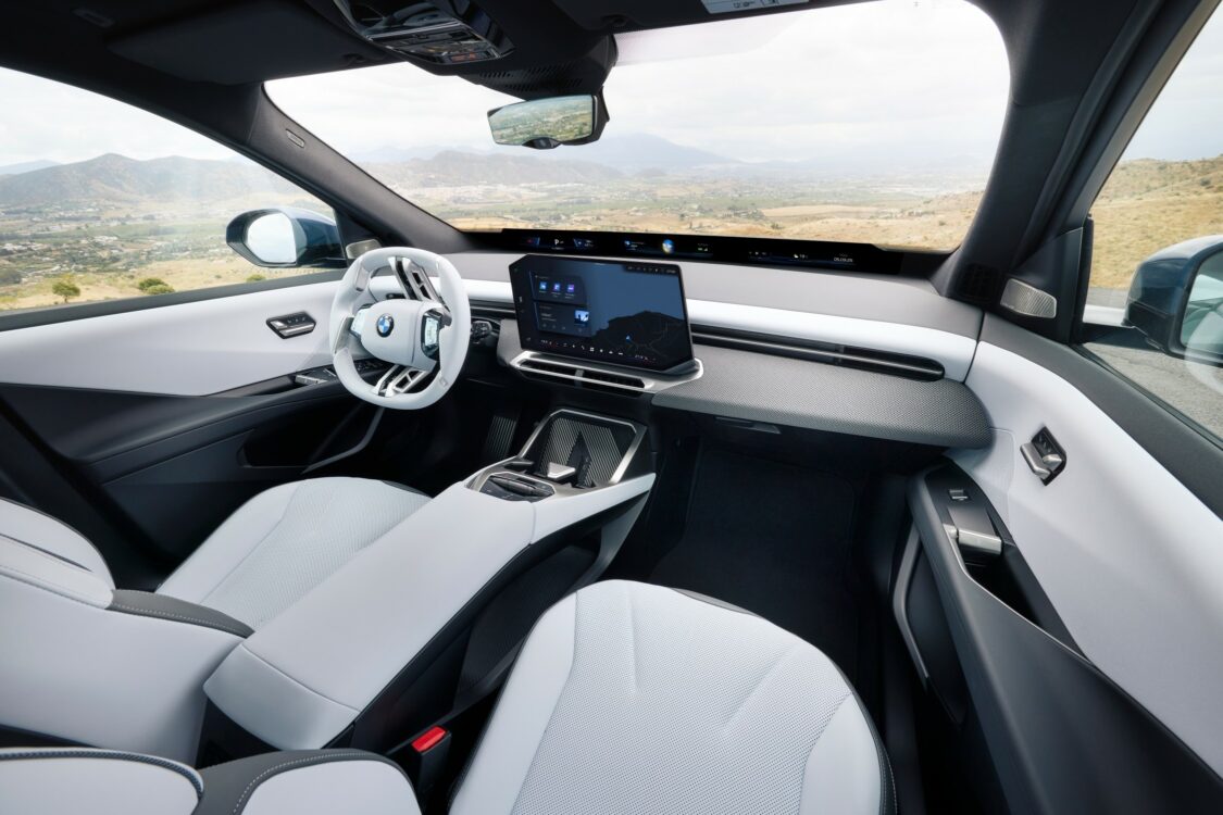 Intérieur BMW iX3 – élégance et technologie face aux montagnes Intérieur de la BMW iX3 vu côté passager, avec sièges blancs, volant blanc et grand écran central, sur fond de paysage montagneux.
