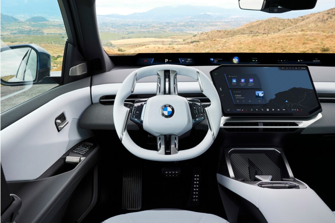 BMW iX3 – cockpit moderne et digital face aux montagnes Gros plan sur le volant blanc et l’écran central de la BMW iX3, avec affichage de navigation, sur fond de paysage montagneux.
