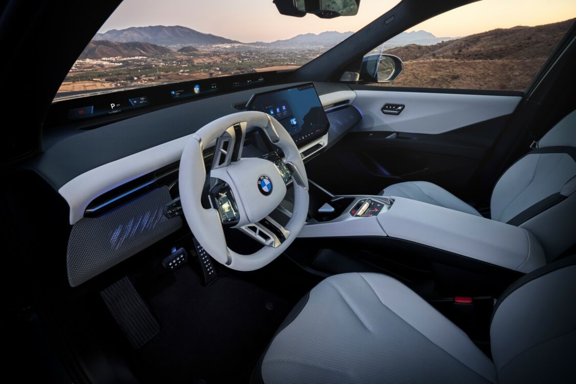 BMW iX3 – design intérieur élégant et technologique Vue oblique de l’intérieur de la BMW iX3, montrant le volant blanc, l’écran central et la console, avec un paysage montagneux en arrière-plan.