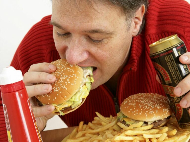Homme mangeant un fast-food gras, avec deux burgers, des frites, une bière et du ketchup.